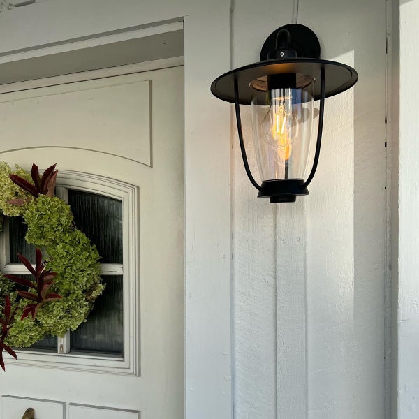 Schwarze Außenwandleuchte im Laternen-Stil aus Metall an einer Wand neben einer Tür mit Kranz, klares Glas mit leuchtender Filamentlampe.