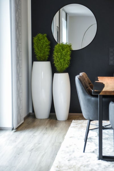 Modernes Esszimmer mit zwei hohen weißen Bodenvasen und Grünpflanzen vor einer dunklen Wand mit einem runden Spiegel.
