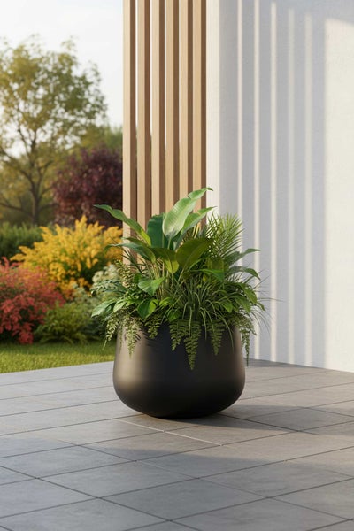 Großes rundes Pflanzgefäß in Schwarz auf einer Terrasse mit verschiedenen grünen Pflanzen vor einer modernen Fassade und einem Garten.