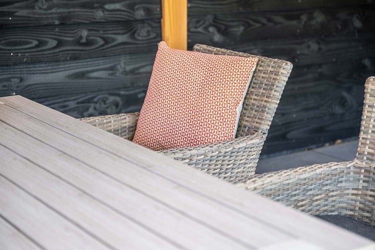 Nahaufnahme einer Garten-Sitzgruppe mit Holztisch und Polyrattan-Sessel sowie einem gemusterten Dekokissen auf einer Terrasse.