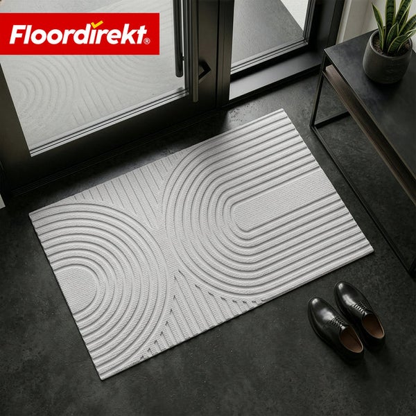 Floordirekt Logo. Rechteckige Fußmatte mit strukturiertem geometrischem Muster aus konzentrischen Bögen, platziert im Eingangsbereich vor Glastür.