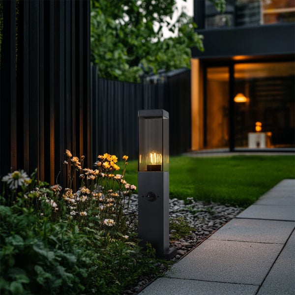 Moderne schwarze Außenwegeleuchte mit Bewegungsmelder und Filament-Leuchtmittel im Garten neben einem Steinweg und Blumenbeet.