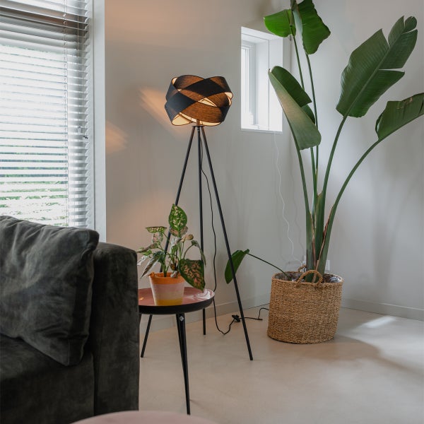 Moderne Wohnzimmerszene mit einer schwarzen Dreibein-Stehleuchte mit geflochtenem Schirm, einer großen Zimmerpflanze im Korb und einem Beistelltisch.