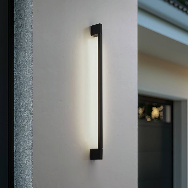 Moderne schwarze LED-Außenwandleuchte in vertikaler Stabform an einer weißen Hauswand mit warmem indirektem Licht.