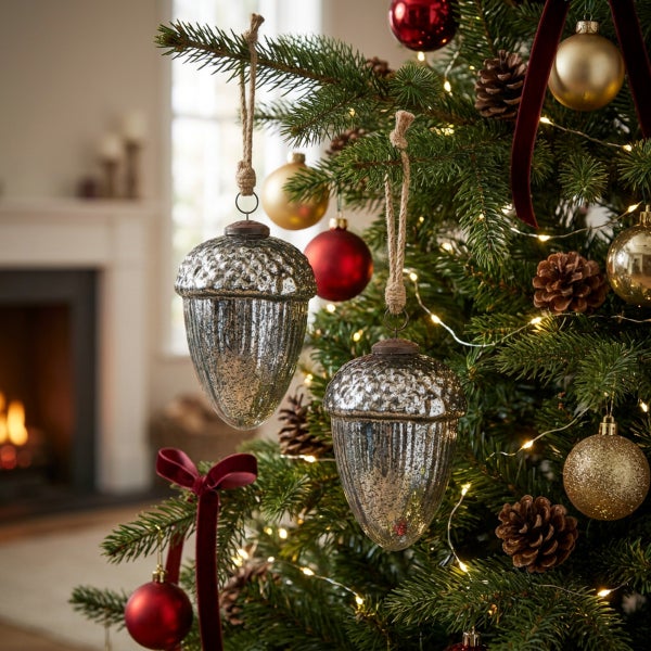 Zwei dekorative Anhänger in Eichelform aus antik wirkendem Glas hängen an Juteschnüren in einem geschmückten Weihnachtsbaum mit Lichterkette.