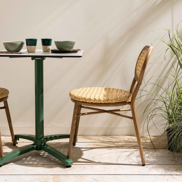 Bistrotisch mit grünem Metallfuß und Stuhl mit Rattangeflecht auf einer hellen Terrasse.