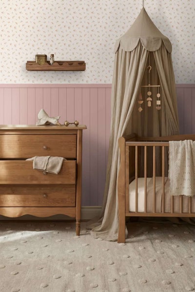 Kinderschlafzimmer mit Holzkommode, Gitterbett mit Betthimmel, Blümchentapete, Wandpaneelen und Wandregal in harmonischer Raumgestaltung.