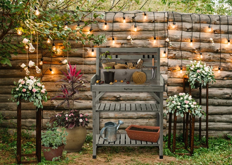 Grauer Pflanztisch aus Holz mit Arbeitsfläche und Ablageflächen, dekoriert mit Gartenwerkzeug, Pflanzen und Lichterkette vor einer Holzwand.