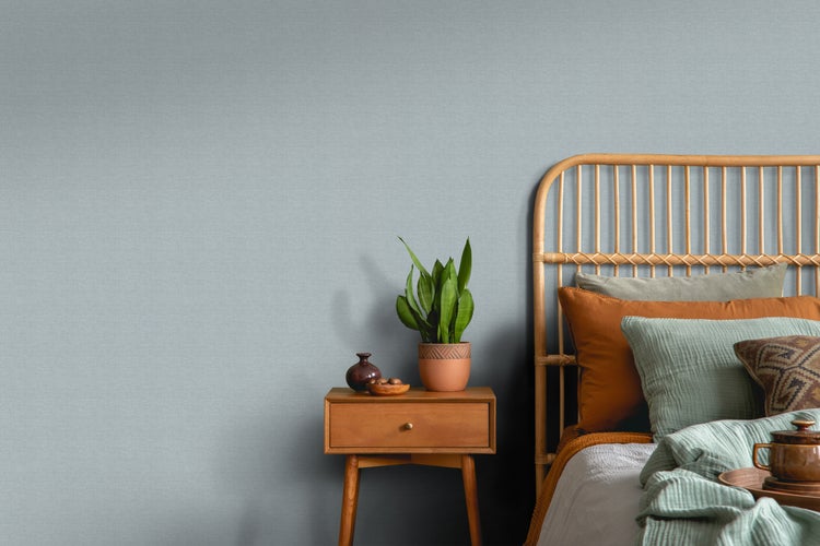 Schlafzimmer mit Rattanbett, Nachttisch und Zimmerpflanze vor einer strukturierten Tapete.