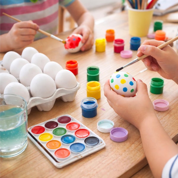 Kinder bemalen Ostereier mit Pinseln und verschiedenen Farben an einem Holztisch mit Farbkasten und Farbtöpfen.