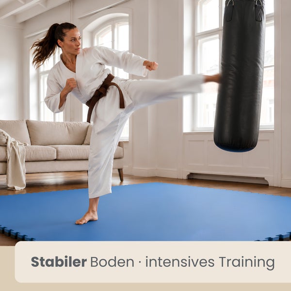 Frau im Karate-Anzug trainiert an einem Boxsack auf blauen Bodenschutzmatten in einem hellen Wohnzimmer.