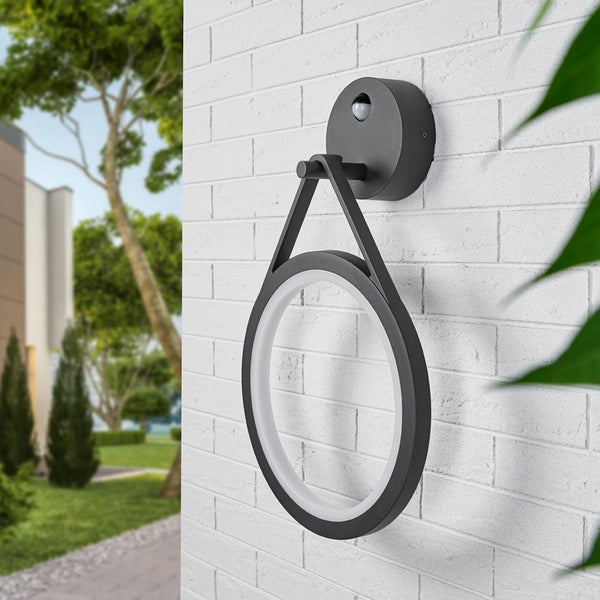 Moderne, ringförmige LED-Außenwandleuchte mit Bewegungsmelder, montiert an einer weißen Ziegelwand in einer hellen Gartenumgebung.