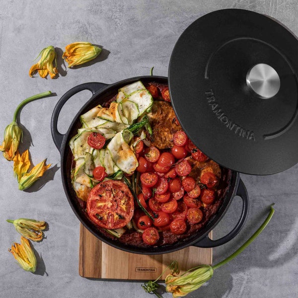 Gusseisenbräter mit Deckel, gefüllt mit Gemüse wie Tomaten und Zucchini, auf einem Holzbrett. Marke Tramontina.