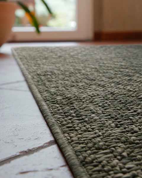 Nahaufnahme eines flachgewebten Bouclé-Teppichs mit strukturierter Oberfläche und geketteltem Rand auf einem Fliesenboden.