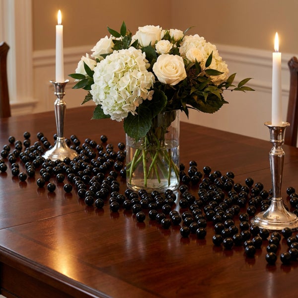 Tischdekoration mit einem Blumenstrauß aus weißen Blumen in einer Glasvase, flankiert von zwei Kerzenhaltern und verstreuten schwarzen Deko Kugeln.