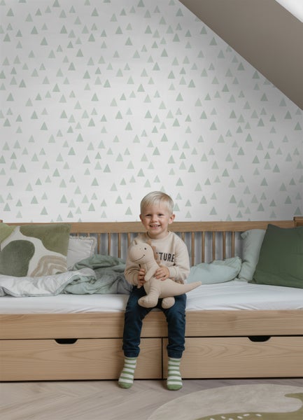 Kinderzimmer mit heller Tapete mit grünem Dreiecksmuster an der Wand, ein Kind sitzt auf einem Holzbett mit Schubladen.