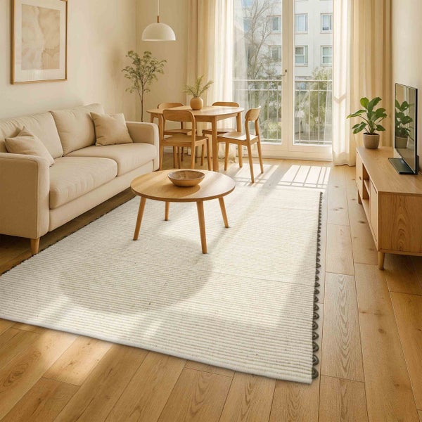 Hell eingerichtetes Wohnzimmer mit cremefarbenem Sofa, rundem Holztisch und großem hellen Teppich auf hellem Holzboden vor Balkontüren.