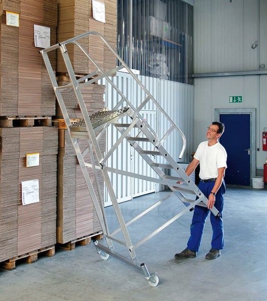 Ein Mitarbeiter in einem Lagerhaus bewegt eine fahrbare Aluminiumtreppe mit Plattform zwischen gestapelten Paletten.