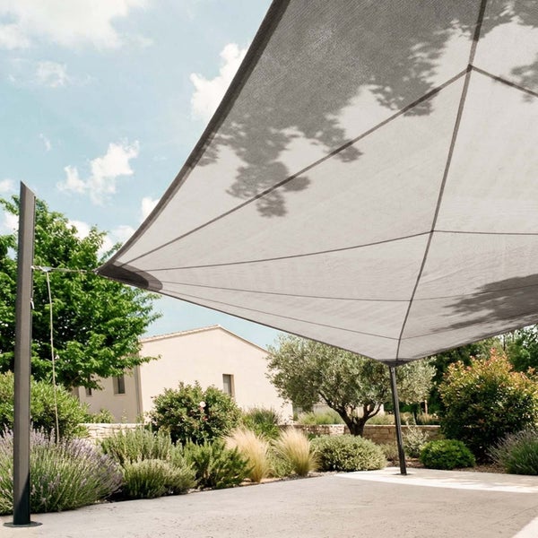 Großes Sonnensegel an Metallmasten über einer hellen Gartenterrasse mit vielfältiger Bepflanzung und einem Wohnhaus im Hintergrund.