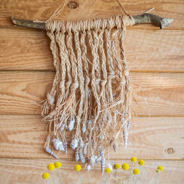 Handgefertigte Wanddekoration aus geflochtenem Naturstroh an einem Holzast, verziert mit kleinen Muscheln.