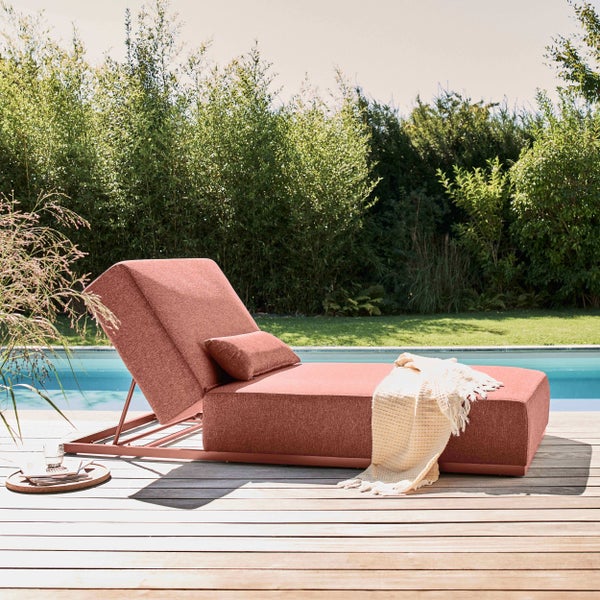 Moderne gepolsterte Sonnenliege auf einer Holzterrasse am Pool, umgeben von grünen Hecken.