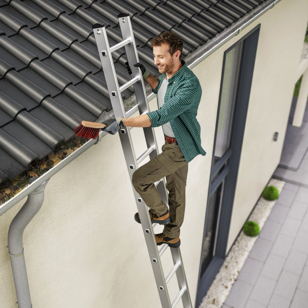Ein Mann auf einer Anlegeleiter reinigt eine Dachrinne mit einer Bürste an einem Haus mit Ziegeldach.