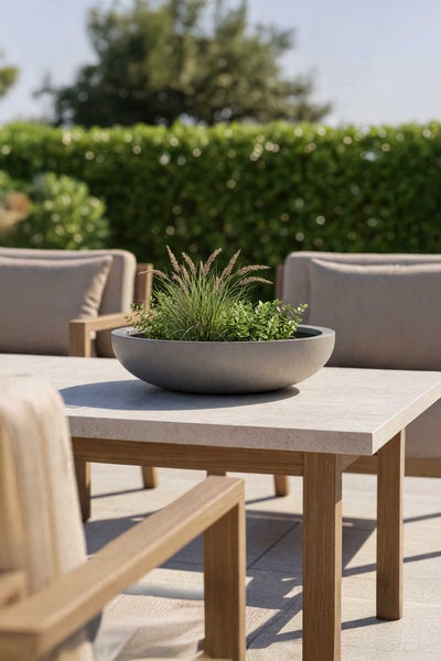 Flache Pflanzschale in Betonoptik mit Gräsern auf einem modernen Gartentisch aus Holz und Stein auf einer hellen Terrasse.
