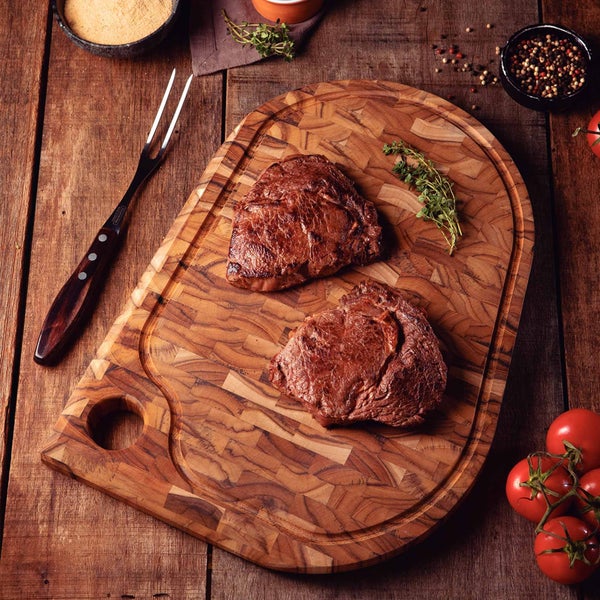 Holzschneidebrett mit zwei Steaks, Gabel, Kräutern und Tomaten auf einem Holztisch.