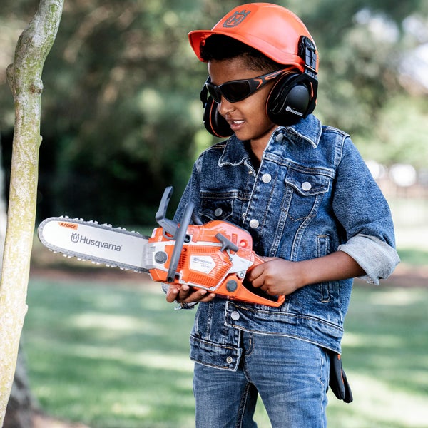 Junge mit Spielzeugkettensäge, Schutzhelm und Schutzbrille im Garten.