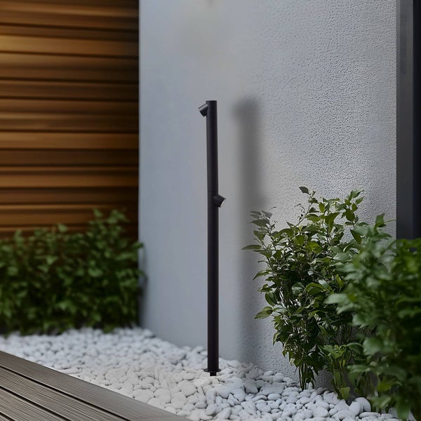 Schwarze Außen-Pollerleuchte mit zwei verstellbaren Strahlern in einem Garten mit weißen Kieselsteinen und grünen Pflanzen vor einer Wand.