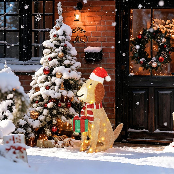 Beleuchtete LED-Hundefigur mit Weihnachtsmütze und Geschenk neben verschneitem Weihnachtsbaum vor einem Hauseingang.