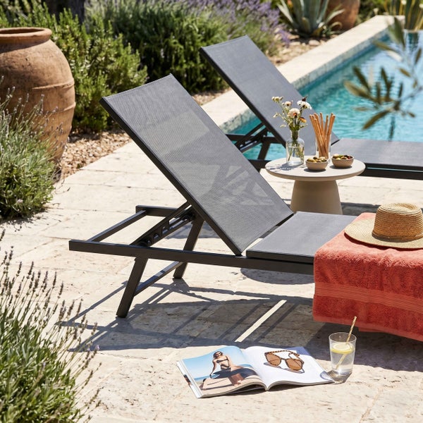 Moderne Gartenliege mit verstellbarer Rückenlehne auf einer hellen Steinterrasse am Pool, umgeben von Pflanzen und Zubehör.