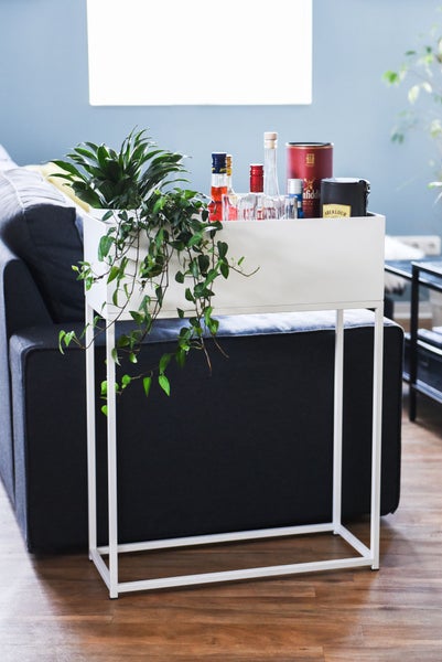 Weißer Pflanzenständer aus Metall als Barwagen mit Getränkeflaschen, Gläsern und Grünpflanze im Wohnzimmer neben einem Sofa.