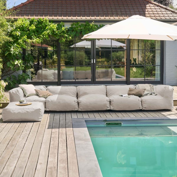 Modulares Outdoor-Sofa-Set auf einer Holzterrasse neben einem Swimmingpool, inklusive Hocker, Kissen und großem Sonnenschirm vor einer Fensterfront.