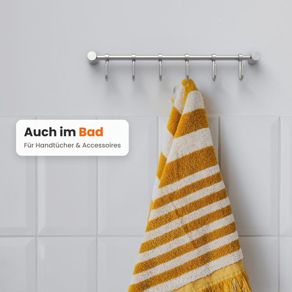 Wandgarderobe mit sechs Haken aus Metall an einer weißen Fliesenwand montiert, mit einem gestreiften Handtuch für die Anwendung im Badezimmer.
