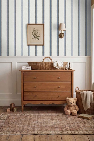 Kindertagesraum mit gestreifter Tapete, einer hölzernen Kommode mit Weidenkorb, einem Teddybär und einem Vintage Teppich auf Holzboden.