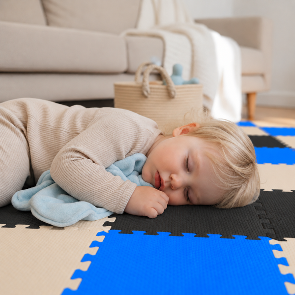 Ein Kleinkind schläft auf einer weichen, zusammensteckbaren Spielmatte aus Schaumstoff mit einem blauen Kuscheltuch im Wohnzimmer.