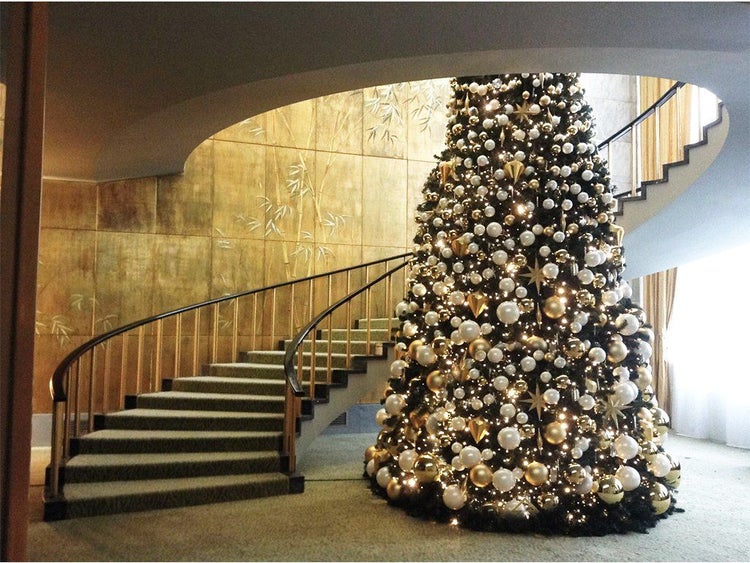 Großer geschmückter Weihnachtsbaum in einem eleganten Treppenhaus mit goldener Wandgestaltung und gewendelter Treppe.