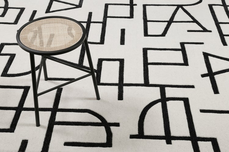 Schwarzer Hocker mit runder Sitzfläche aus Wiener Geflecht auf einem Teppich mit geometrischem Linienmuster.