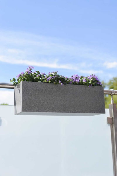 Rechteckiger Balkon-Blumenkasten in Steinoptik, bepflanzt mit lila Blumen, montiert an einem modernen Balkongeländer aus Metall und Glas.