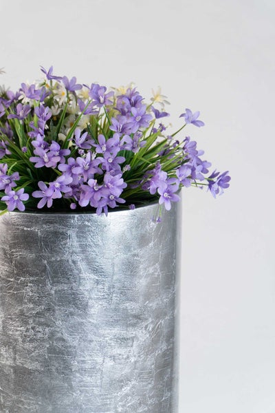 Künstliche Blumen mit violetten Blüten in einem silbernen, strukturierten zylindrischen Übertopf.