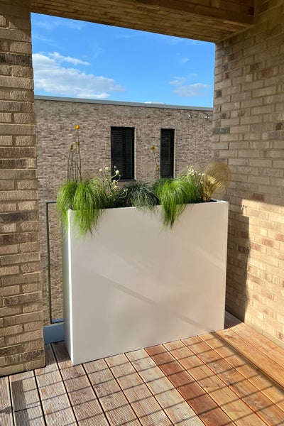 Großer weißer rechteckiger Pflanzkübel auf einem Balkon mit Holzdiele, bepflanzt mit Gräsern und kleinen Blumen in heller, moderner Umgebung.