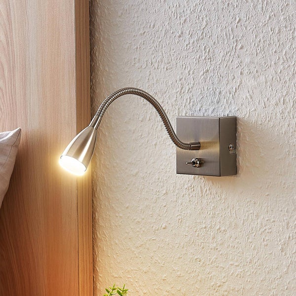 Flexible Wandleuchte aus Metall mit Kippschalter, montiert an einer Wand neben einem hölzernen Kopfteil, Licht ist eingeschaltet.