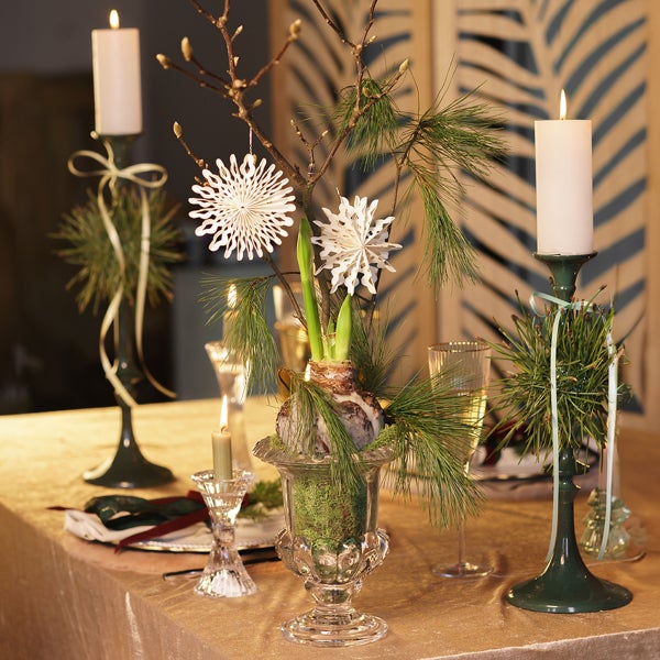 Festlich gedeckter Tisch mit Amaryllis-Gesteck in Glasvase, Kiefernzweigen, Papierschneeflocken und weißen Stumpenkerzen auf grünen Kerzenhaltern.