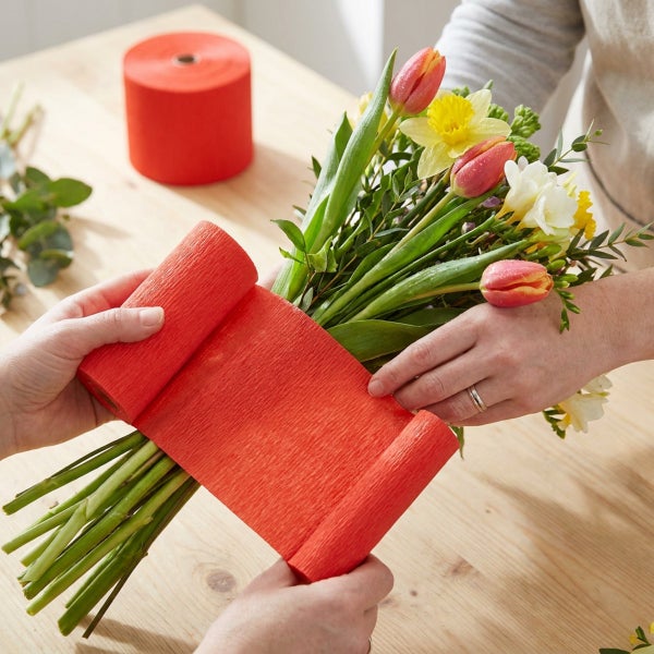 Person wickelt einen Blumenstrauß aus Tulpen und Narzissen in rotes Krepppapier ein.
