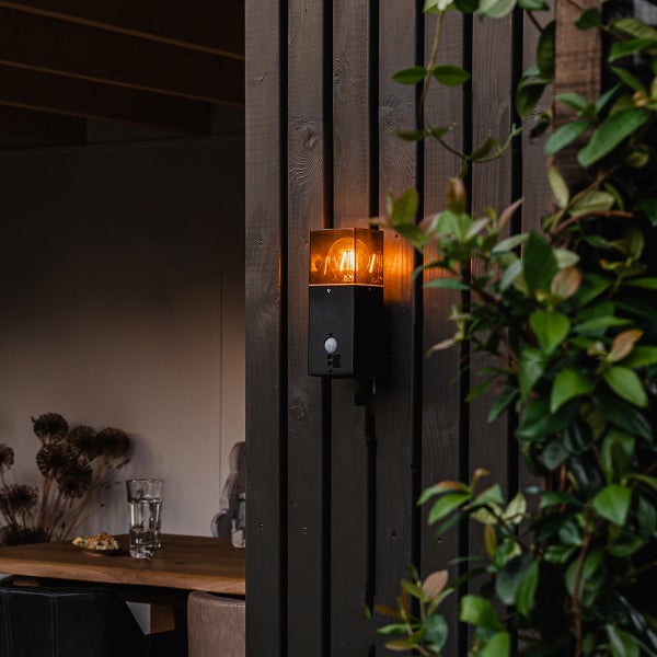 Schwarze Außenwandleuchte mit Bewegungsmelder und warmem LED-Licht an einer Holzwand neben grünen Pflanzen.