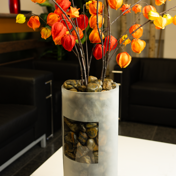 Zylindrische Vase aus mattiertem Glas, gefüllt mit Natursteinen und getrockneten Zweigen der Lampionblume in einem modern eingerichteten Raum.