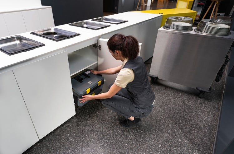 Eine Person kniet vor einem weißen Unterschrank und verstaut einen Werkzeugkoffer aus Kunststoff in einer hellen, organisierten Profiküche.