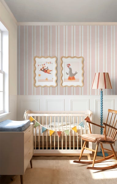 Hell eingerichtetes Kinderzimmer mit vertikal gestreifter Tapete in Rosa und Beige, weißem Gitterbett, Holzschaukelstuhl und Wandbildern.