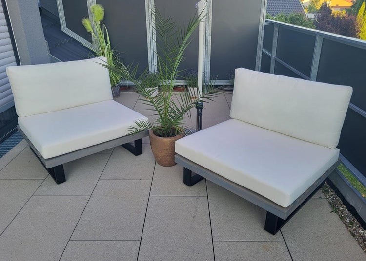 Zwei moderne Garten-Lounge-Sessel mit weißen Polstern und eine Topfpflanze auf einer hellen Terrasse mit Steinboden.
